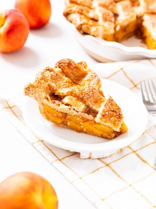 White plate with slice of peach pie sitting amongst the rest of the pie with a few peaches scattered around.
