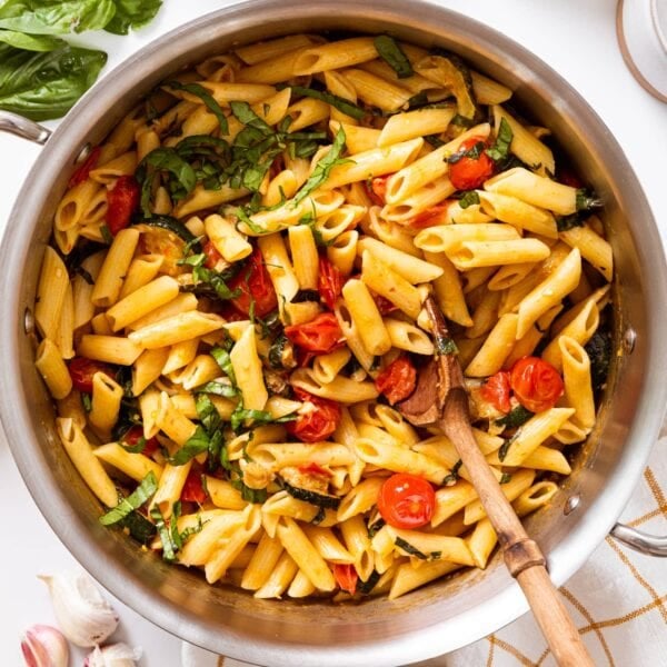Metal stockpot sitting on white countertop filled with penne pasta along with tomatoes and zucchini and basil pieces.