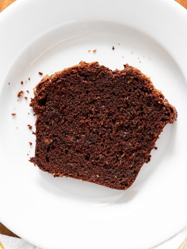 Slice of brown chocolate zucchini bread sitting on white plate with crumbs all around.