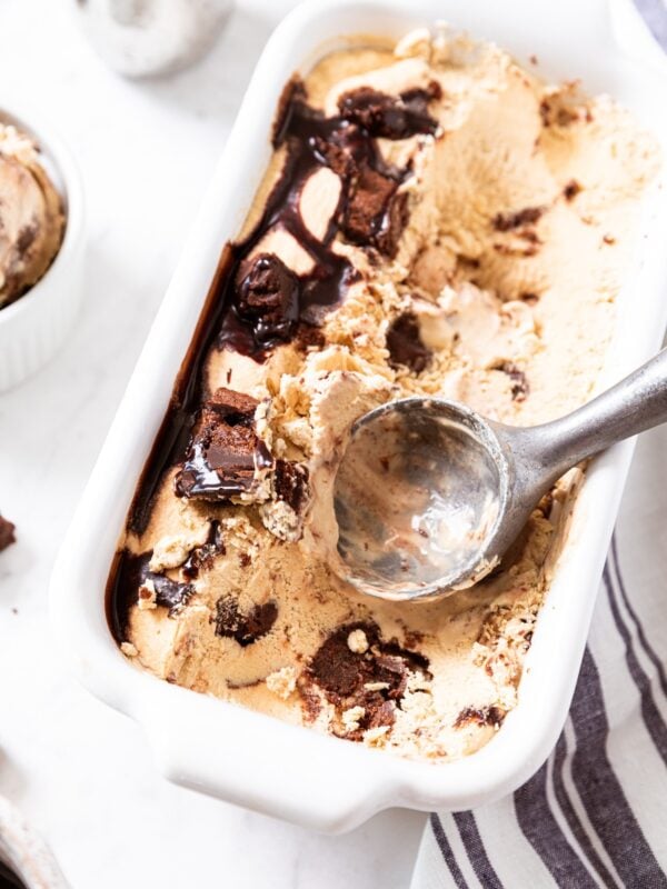 White loaf pan with metal ice cream scoop resting in espresso brownie ice cream on white surface.