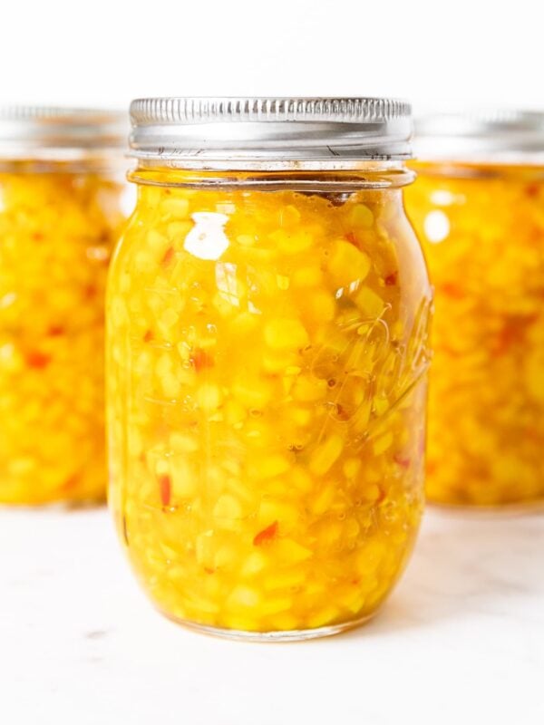 Glass jar sitting on white countertop filled with yellow colored corn relish with red specks of bell pepper inside.
