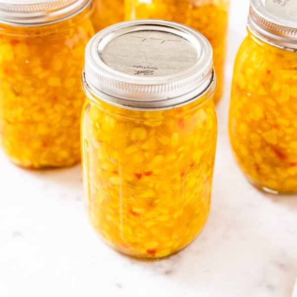White marble countertop filled with glass jars with lids and rings on top filled with yellow colored corn relish with red specks.