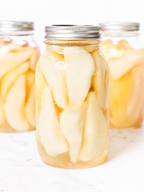 Three glass canning jars filled with sliced canned pears sitting on white marble surface with lids and rings on top.