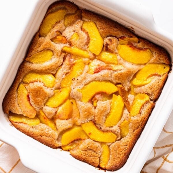 White baking dish filled with baked lazy peach cobbler with large peach slices showing on top of cobbler sitting on white countertop.