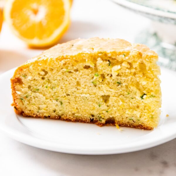 Slice of olive oil cake sitting on white plate on marble surface with orange in background.