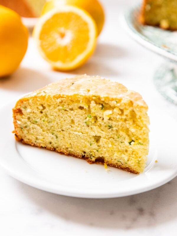 Slice of yellow colored olive oil cake sitting on white plate with orange slice and rest of cake in background all on marble surface.