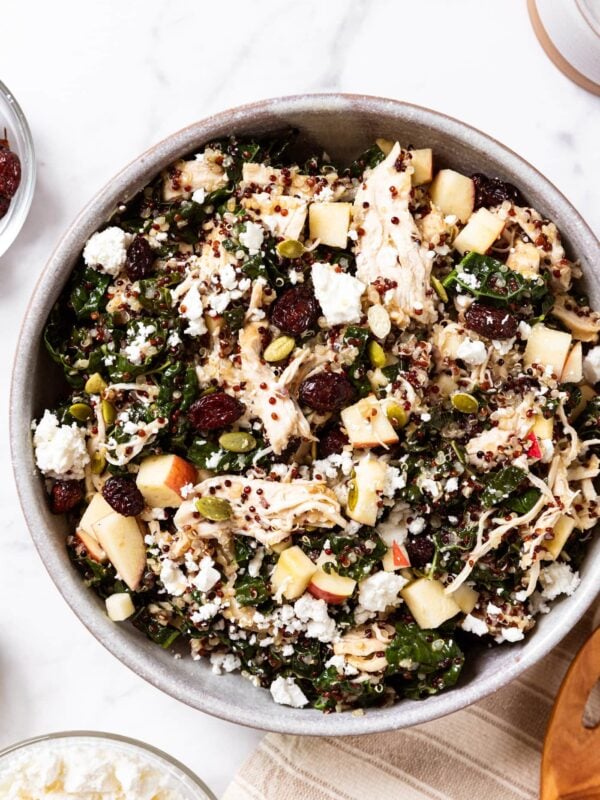 Marble surface with large salad bowl filled with kale salad topped with shreds of chicken, pumpkin seeds, feta cheese, and dried cranberries.