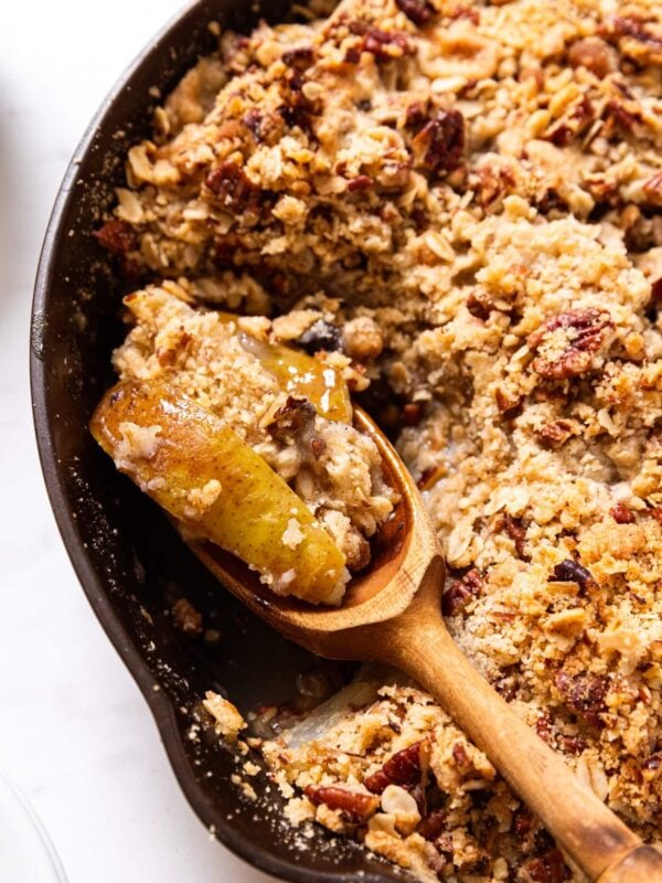 Black skillet filled with pear skillet crisp with wooden spoon scooping up serving with one baked pear on it all on white surface.