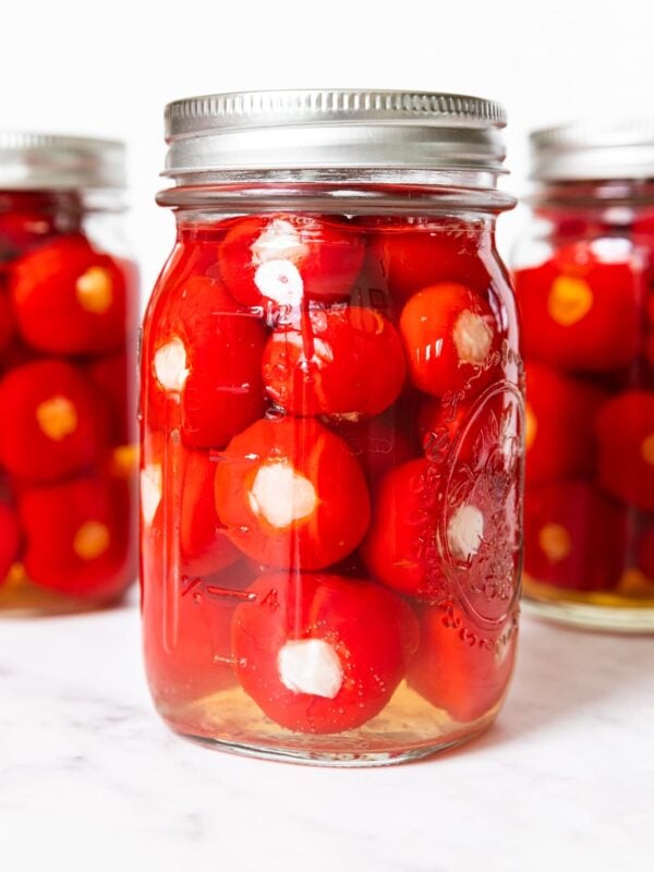 Glass jars filled with red colored miniature peppers stuffed with cabbage all in yellow colored brine on marble surface.