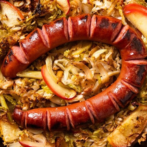 Large black cast iron skillet filled with cooked cabbage and apple with browned piece of sliced kielbasa on top all on marble surface.