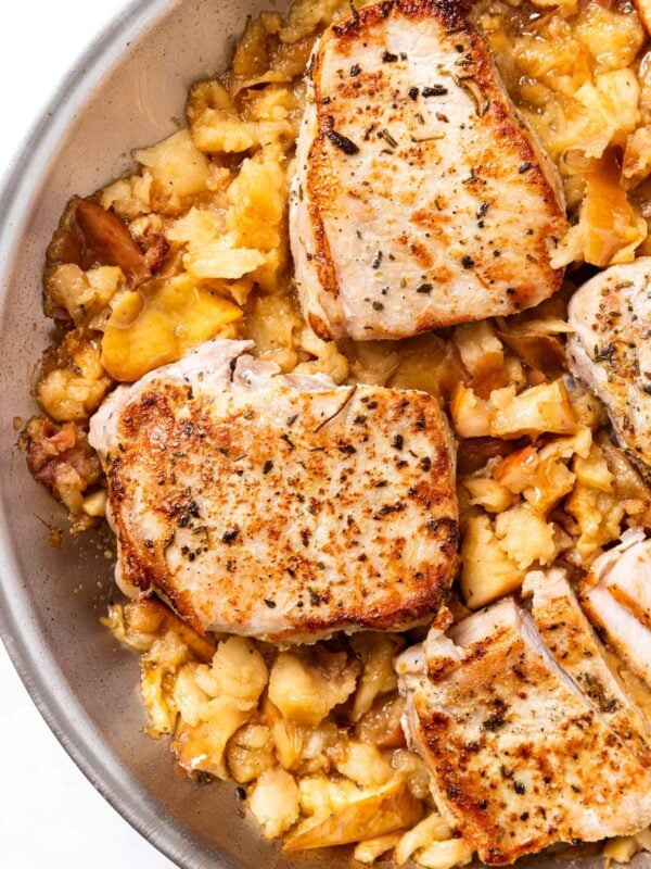 Metal skillet filled with mashed apples and topped with browned pork chops with herbs on top sitting on marble surface.