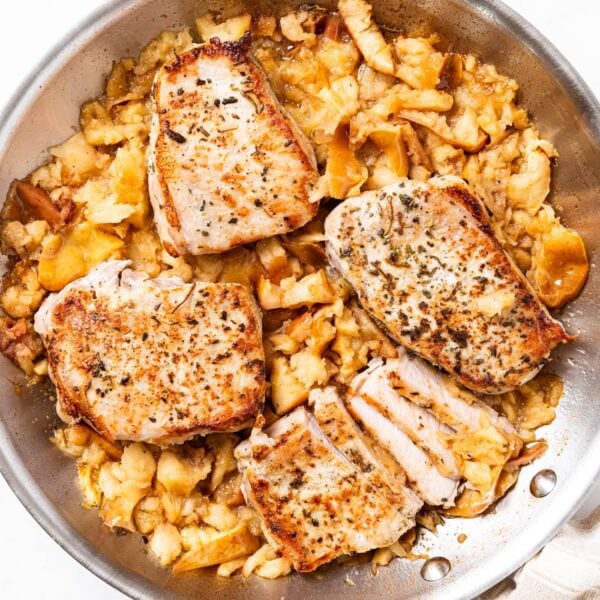 Large metal skillet sitting on white marble with cooked pork chops and mashed apples underneath right after coming out of the oven.