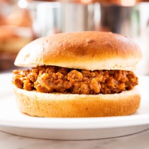 Sloppy Joe meat sitting in between a bun on a white plate with metal pot and copper pots in background.