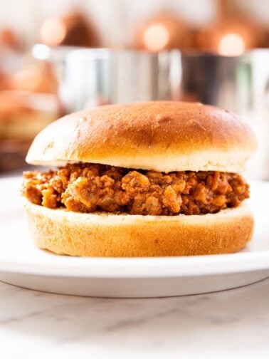 Sloppy Joe meat sitting in between a bun on a white plate with metal pot and copper pots in background.