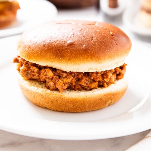 White plate with sandwich bun filled with brown colored sloppy joe meat with top bun on top with more buns and sloppy joe meat in background.