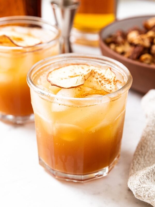 Glass filled with brown colored spiked apple cider sitting on marble surface with bowl full of nuts in background as well as more alcohol needed to make drinks.