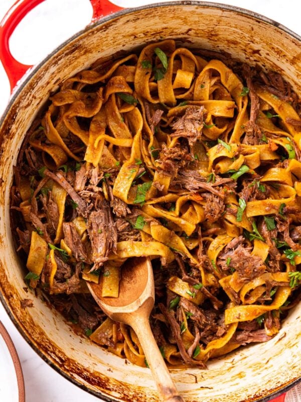 Cooked beef and noodles sitting in red Dutch oven with wooden spoon resting on bottom of pan all on marble surface.