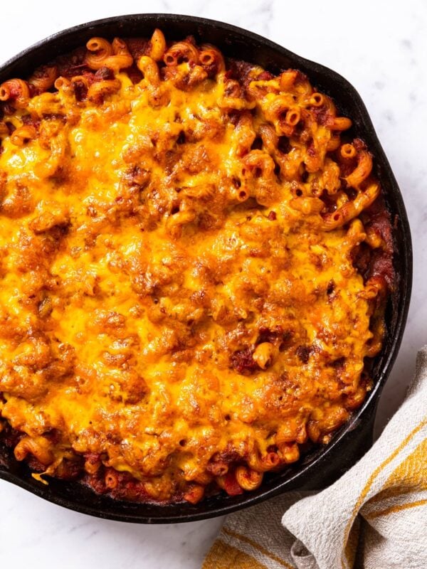 Black cast iron skillet with baked goulash with sprinkled cheese over the top sitting on marble countertop with towel around handle of skillet right after coming out of the oven.