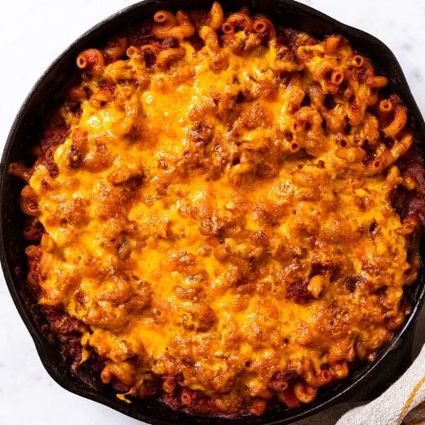 Black cast iron skillet with homemade goulash baked inside sprinkled with cheddar cheese on top with towel covering handle of skillet.