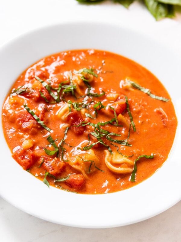 White bowl filled with serving of tomato tortellini soup with chopped basil and Parmesan cheese on top.