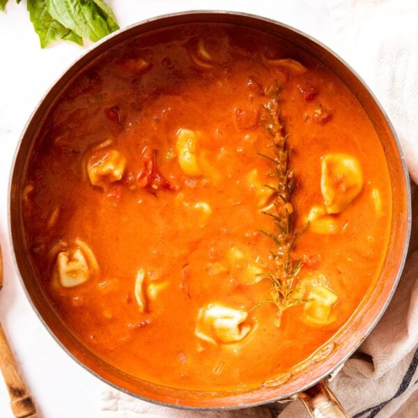 Marble surface with copper pot filled with tomato tortellini soup with wooden spoon beside and cooked sprig of rosemary on top of soup surface.