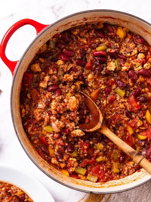 Red Dutch oven sitting on marble surface with wooden spoon resting on top of chili with white bowl filled with more chili to the side.