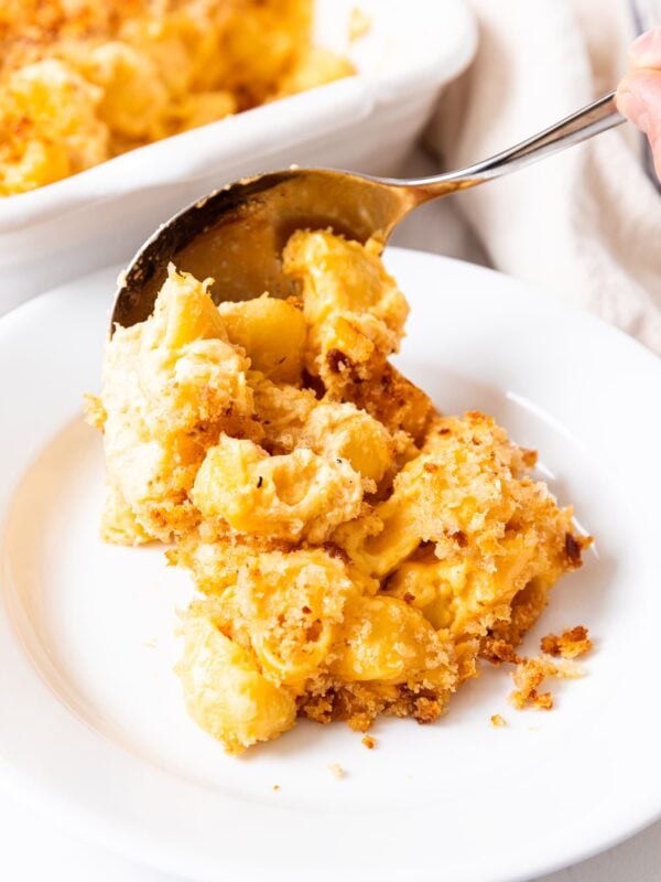 Spoon scooping a serving of macaroni & cheese onto white plate with crumbs escaping and sitting on side of white plate with more casserole in background in white dish.