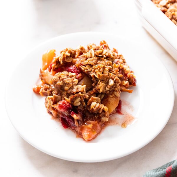 White plate sitting on white marble surface filled with scoop of cranberry crisp with rest of crisp in white baking dish to the side.