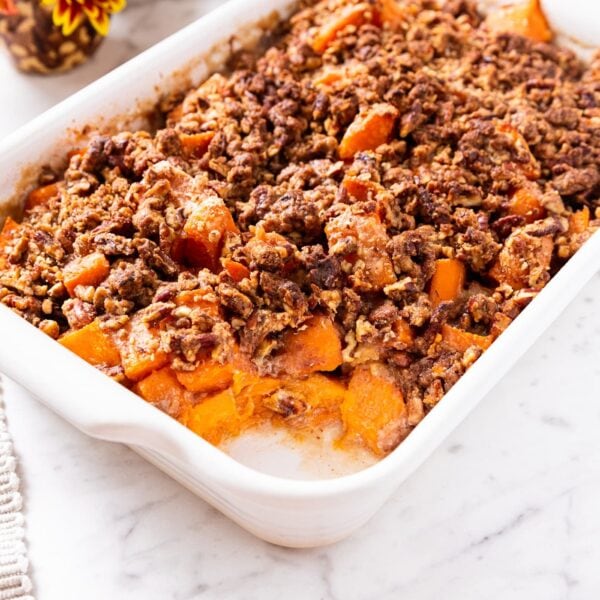 White baking dish filled with sweet potato casserole with pecan streusel topping sitting on marble surface with plate and placemat to the side.
