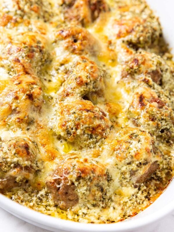 Oval white baking dish filled with baked meatballs with cheese and pesto on top all on white marble surface.