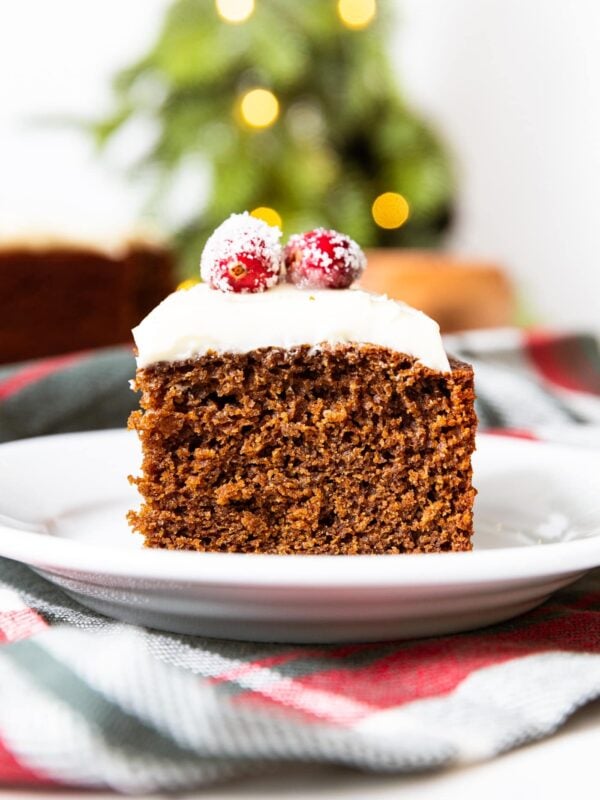 Square of brown colored gingerbread spice cake sitting on with plate with cream cheese frosting and sugared cranberries on top with twinkly lights in the background.