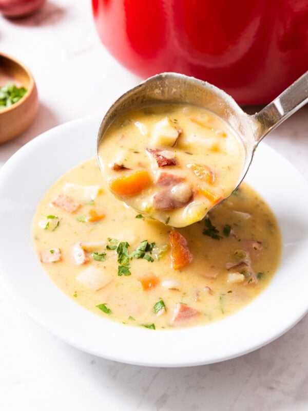 White bowl with scoop of ham and potato chowder being poured on top of soup already in the bowl with red stockpot in the background all on marble surface.