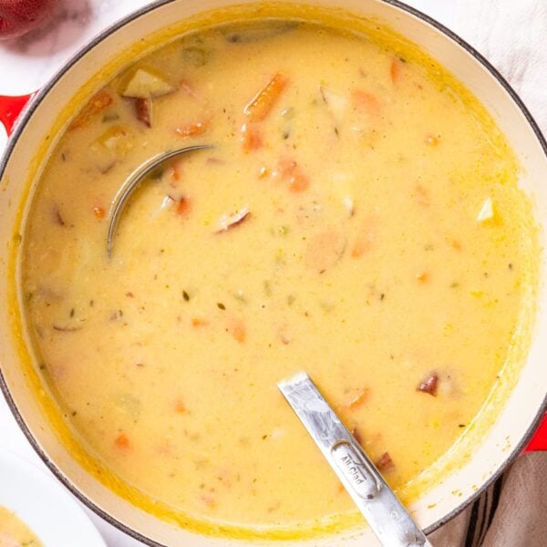 Red stockpot filled with yellow colored ham and potato soup with ladle resting in the bottom with pieces of ham and potato showing through the liquid all on marble surface.
