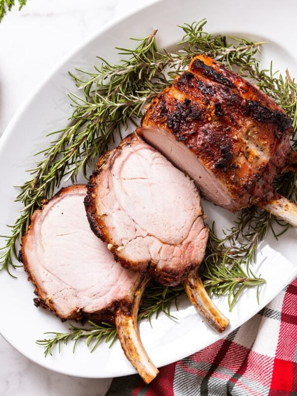 White oval platter filled with roasted pork rib roast with crusty skin with sprigs of fresh rosemary underneath the meat with red and green Christmas napkin underneath the platter.