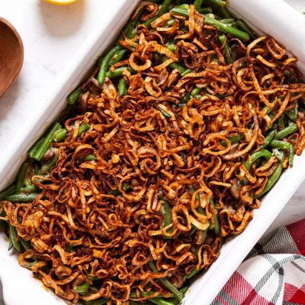 White baking dish filled with baked green bean casserole topped with crispy fried scallions with wooden spoon, lemon, and Christmas napkin to the side all on marble surface.
