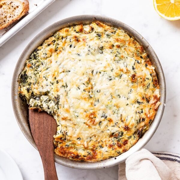 Metal skillet filled with baked spinach artichoke dip with brown top sitting on white marble surface with lemons, Parmesan, and crostini around.