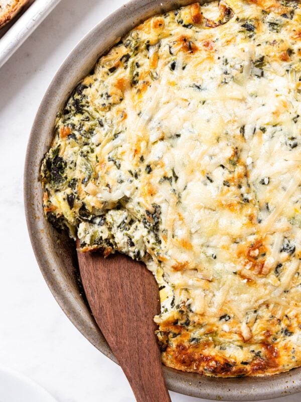 Metal skillet filled with baked spinach artichoke dip with golden top with wooden spoon scooping out a serving of the dip all on white marble surface.