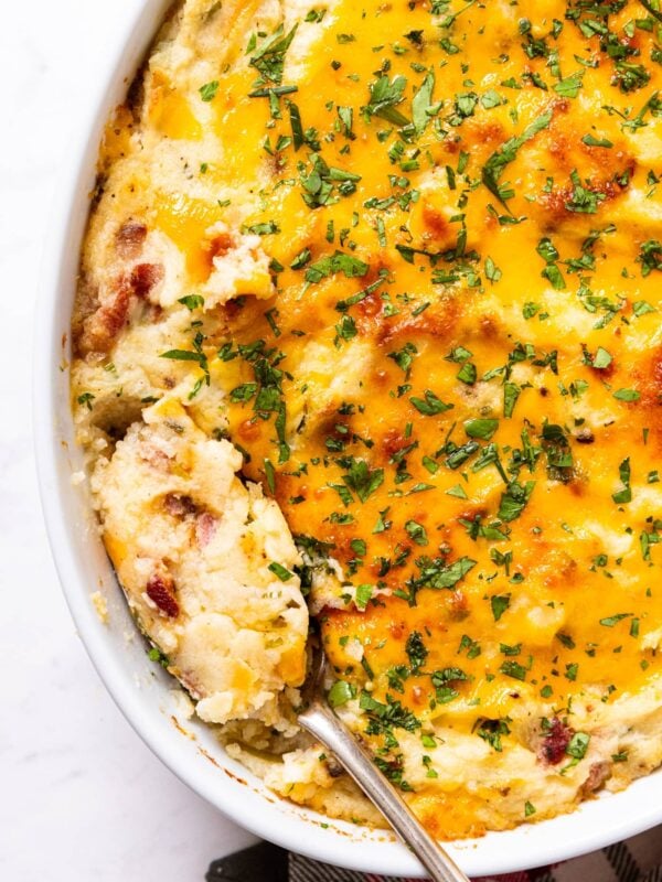 White baking dish filled with twice baked potato casserole with spoon holding a serving of potatoes showing bacon and chives sprinkled on top.