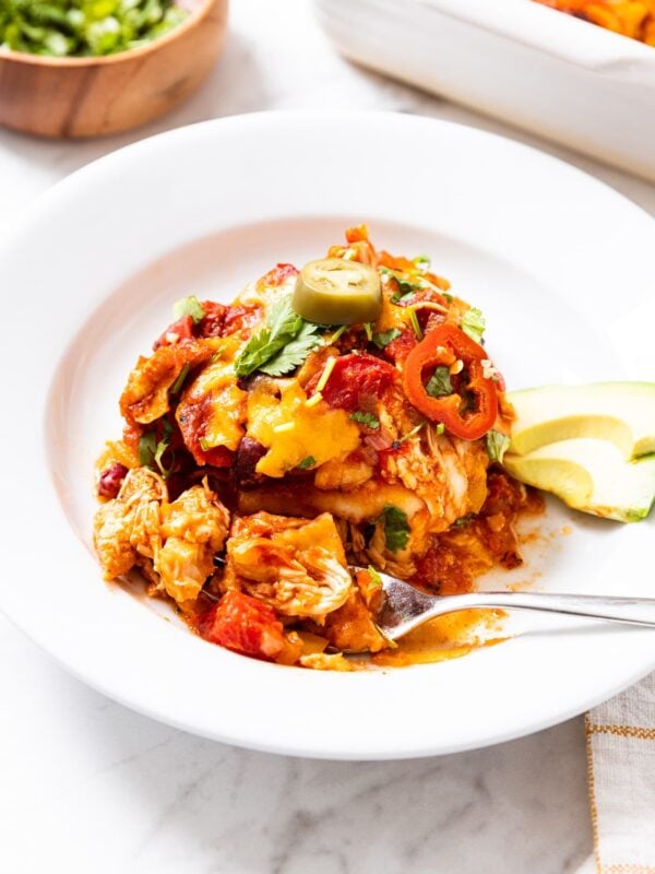 White plate filled with serving of chicken tortilla casserole topped with avocado, cilantro, and spicy peppers with bowl of cilantro in background along with rest of casserole in white baking dish.