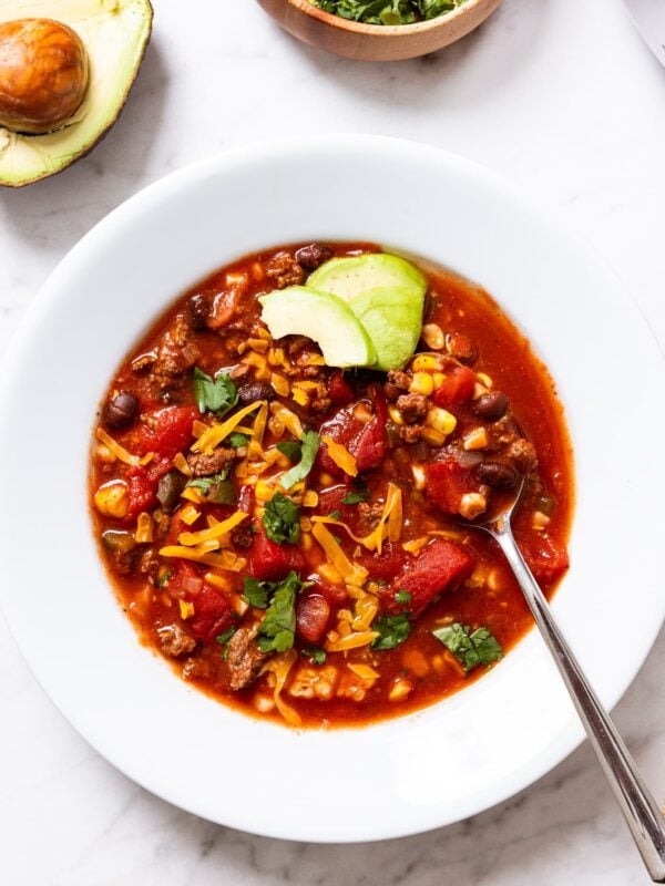 White bowl filled with red colored taco soup showing chunks of tomato, corn, and black beans, with slices of avocado resting on top of soup with spoon resting and ready for first bite.