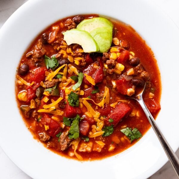 White serving bowl filled with single serving of taco soup topped with cheese and slices of green avocado with spoon resting in bowl all on white marble surface.