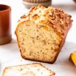 Loaf of classic banana bread sitting on white marble surface with a few slices already sliced off with orange mug filled with coffee to the side along with yellow napkin.