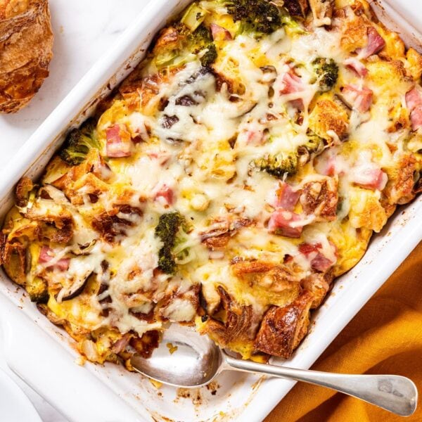 White marble surface with white casserole dish filled with baked ham & cheese croissant egg casserole with cheese sprinkled over the top with spoon sitting in container after scooping out a serving.