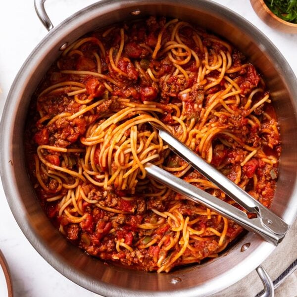Large metal stockpot filled with meat sauce with spaghetti with tongs sitting on top with swirl of noodles all on marble surface with towel, green herbs, and Parmesan cheese.