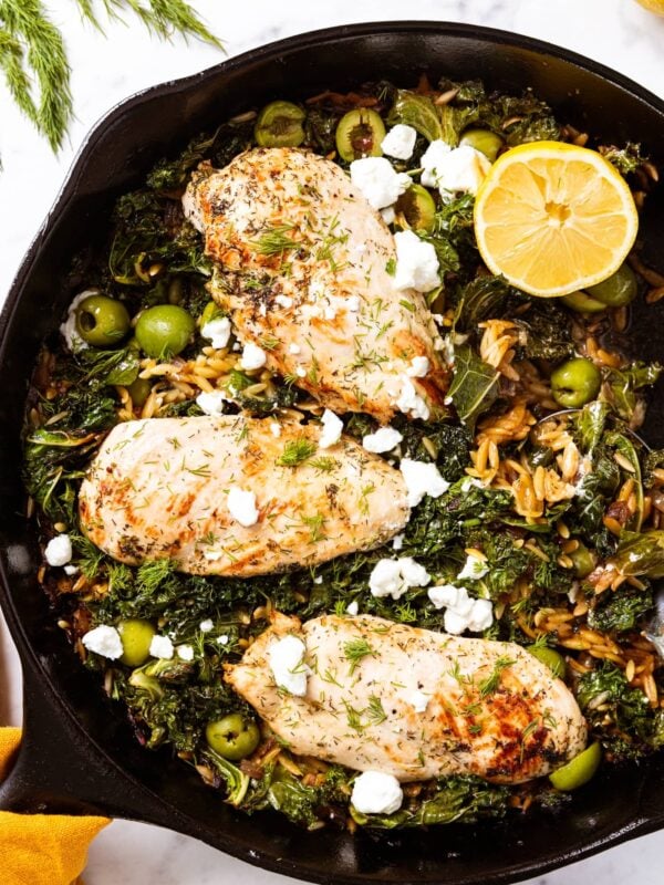 White marble surface with cast iron skillet filled with cooked chicken breasts sitting on top of wilted kale and orzo with crumbled feta cheese on top and a half lemon to the side.