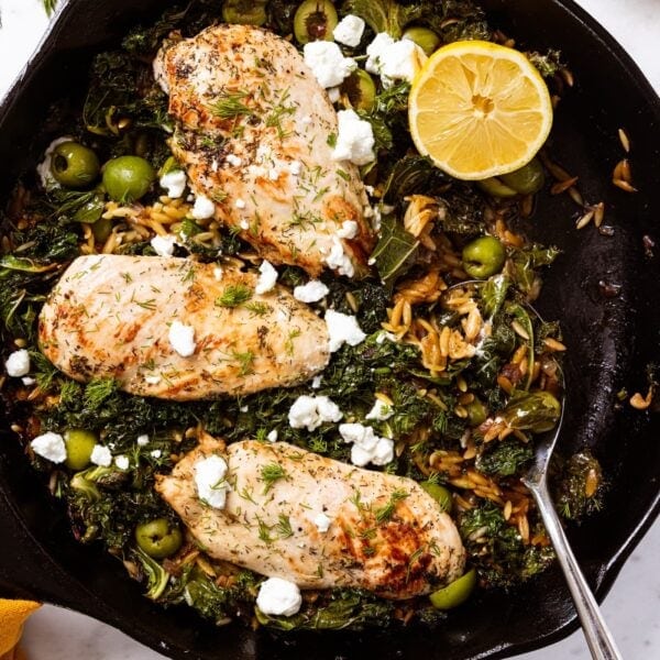 Black cast iron skillet sitting on marble surface filled with cooked chicken breasts and orzo with half of lemon nestled amongst wilted kale with spoon resting on side of skillet.
