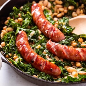 Black skillet filled with wilted kale, white beans, and browned sausage sitting on marble surface with wooden spoon resting on side with Parmesan cheese and salt cellar in background.