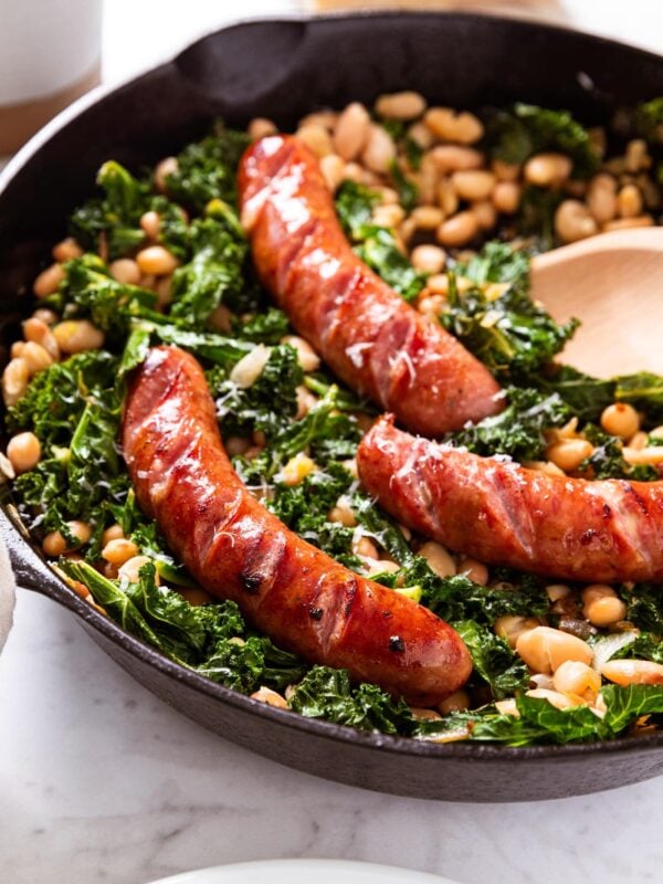 Black skillet filled with wilted kale, white beans, and browned sausage sitting on marble surface with wooden spoon resting on side with Parmesan cheese and salt cellar in background.