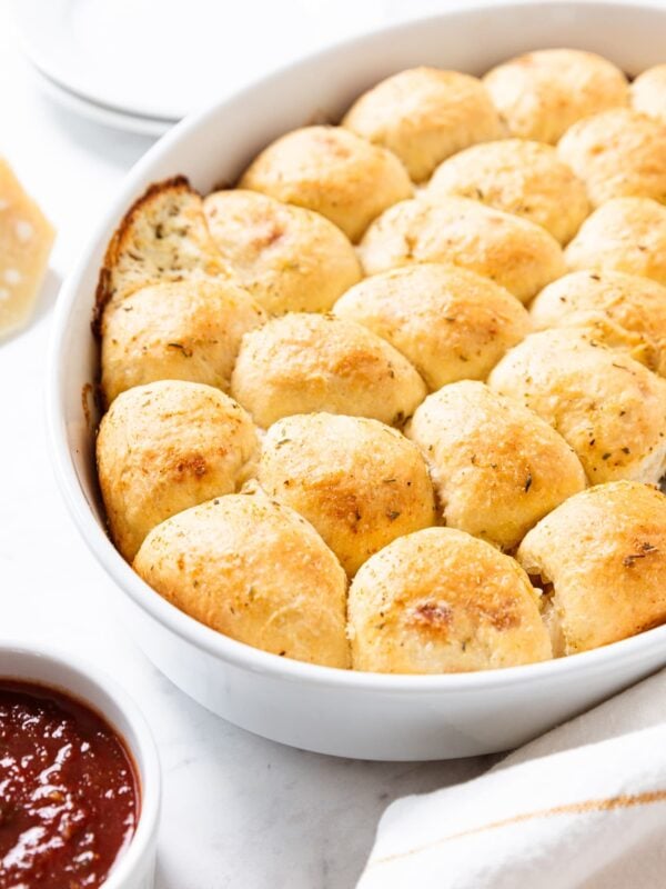 Baked cheese-stuffed pizza bites sitting in oval white baking dish with dish of marinara dipping sauce with white plates in background.