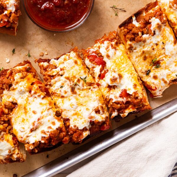 Small baking sheet lined with brown-colored parchment with slices of sausage French bread with small glass bowl filled with tomato dipping sauce.
